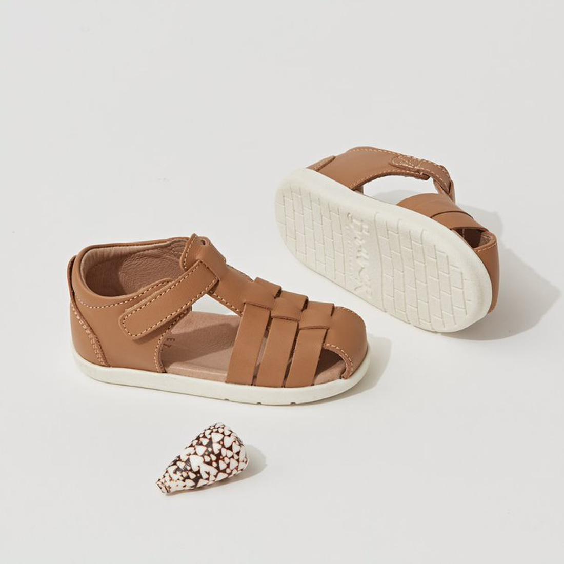 The Pretty Braver Billie First Walker Sandal Closed Back in tan is displayed on a white surface next to a cone-shaped spotted seashell., 9420075334359, 9420075334342, 9421036357264