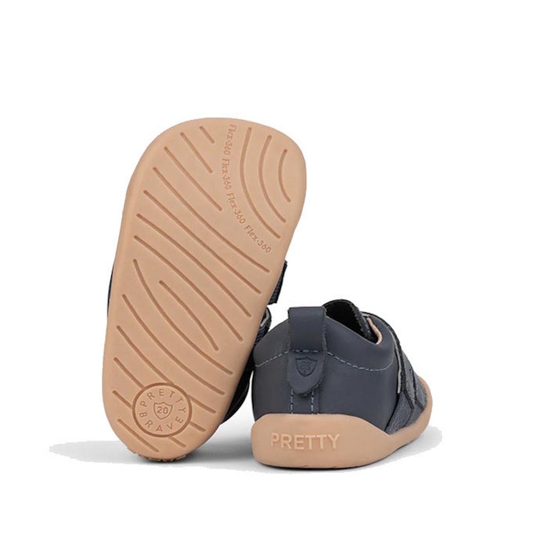 A pair of Pretty Brave First Walker Toto Leather Trainer sneakers in indigo blue leather with light tan flexible rubber soles, featuring curved grooves and "PRETTY BRAVE" stamped on the sole; one shoe stands upright, the other shows the heel., 94200753662