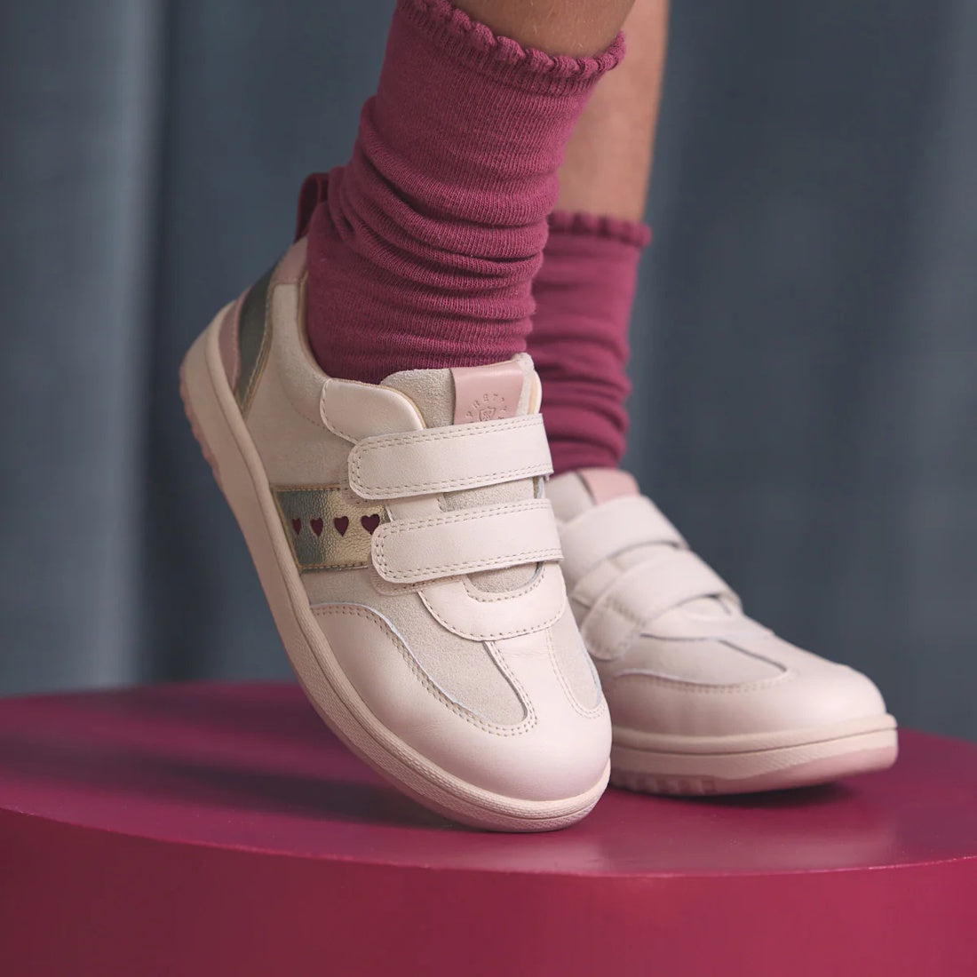 A child wears Pretty Brave River Hearts Leather Trainer Pink Off White with pink socks, standing on a round pink platform against a soft blue background. The shoes feature white genuine leather and small heart designs., 9420075363854, 9420075363847, 94200