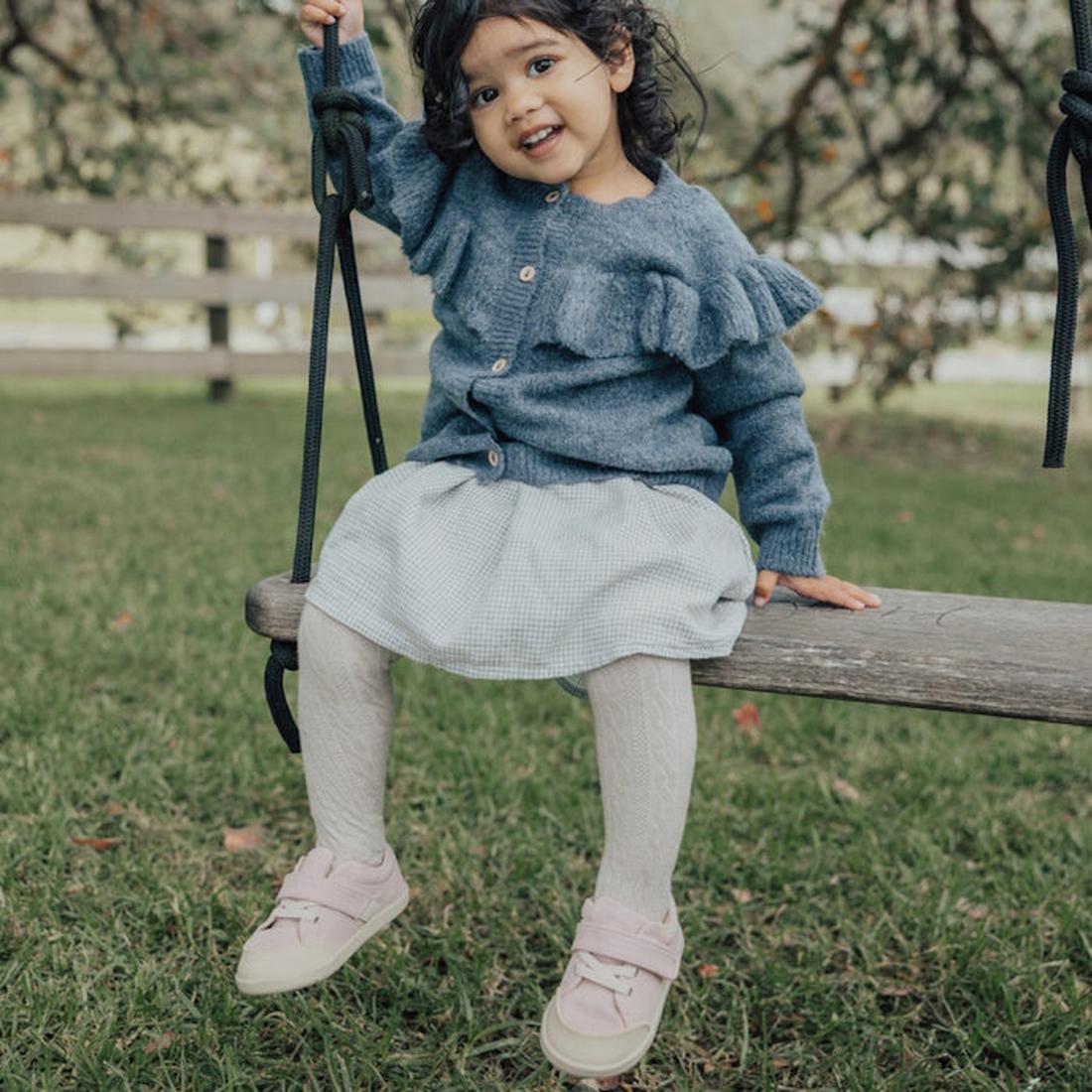 A young child with dark hair smiles while sitting on a wooden swing outside, wearing a blue sweater, light skirt, white tights, and Pretty Brave Boston CANVAS Trainer First Walker Blush Pink shoes. Green grass and a wooden fence are in the background., 94
