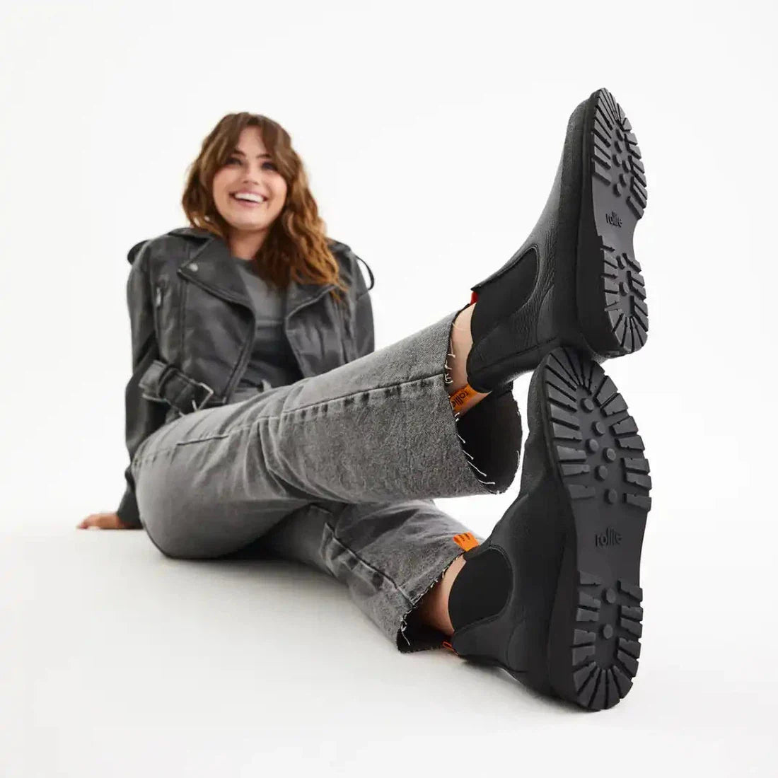A cheerful woman sits cross-legged in grey jeans and a black jacket, highlighting Rollie Womens Fields Chelsea boots’ black color, waterproof finish, easy slip-on style, and rugged soles—ideal for busy family days.  ksnz