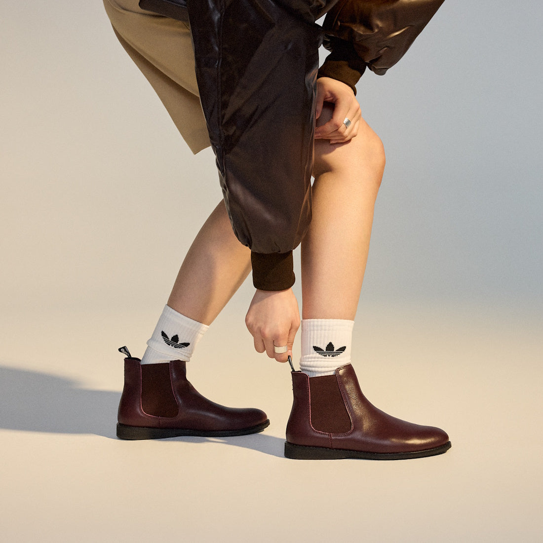 A person wearing Rollie Womens Chelsea Dark Cherry Leather Ankle Boots and white socks with black Adidas logos bends down to adjust one boot. Only the lower body is visible, styled with a brown skirt and shiny brown jacket (SC02136 look)., 9339554019543, 