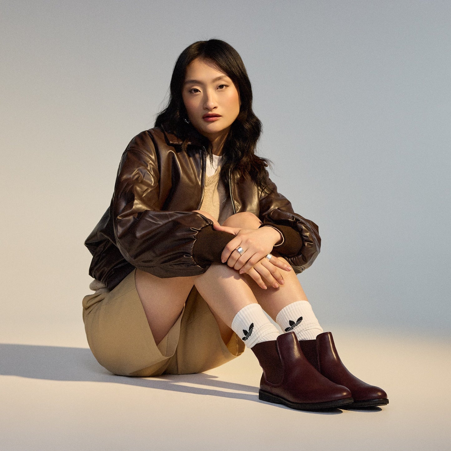 A woman with long dark hair sits on the floor, knees up, looking at the camera. She wears a brown leather jacket, beige shorts, SC02136 socks with black leaf logos, and Rollie Womens Chelsea Dark Cherry Leather Ankle Boot., 9339554019543, 9339554104898, 9