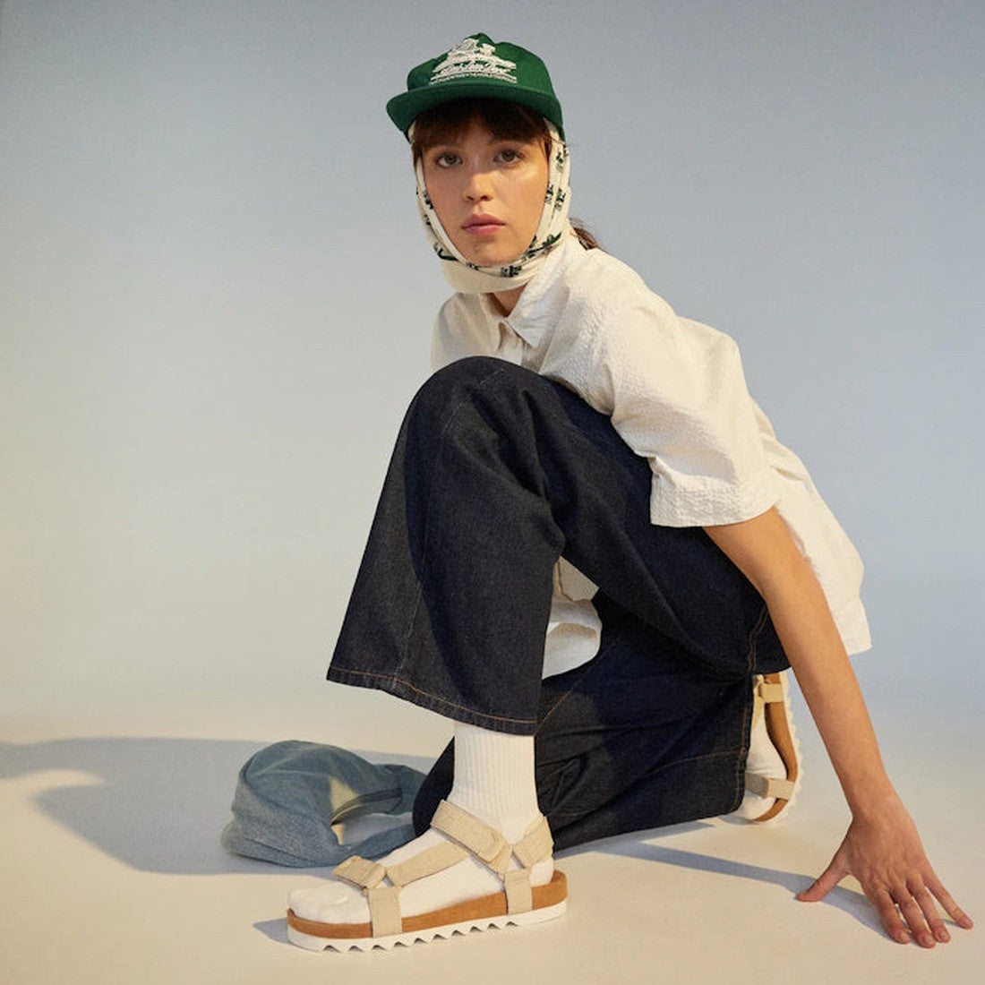 A person wearing a white shirt, wide-leg dark jeans, and Rollie Womens Sandal Tooth Wedge Bone Croc with socks—featuring a suede-wrapped footbed and adjustable sheep leather straps—squats on one knee against a plain background., 9781760775865, 97817607758