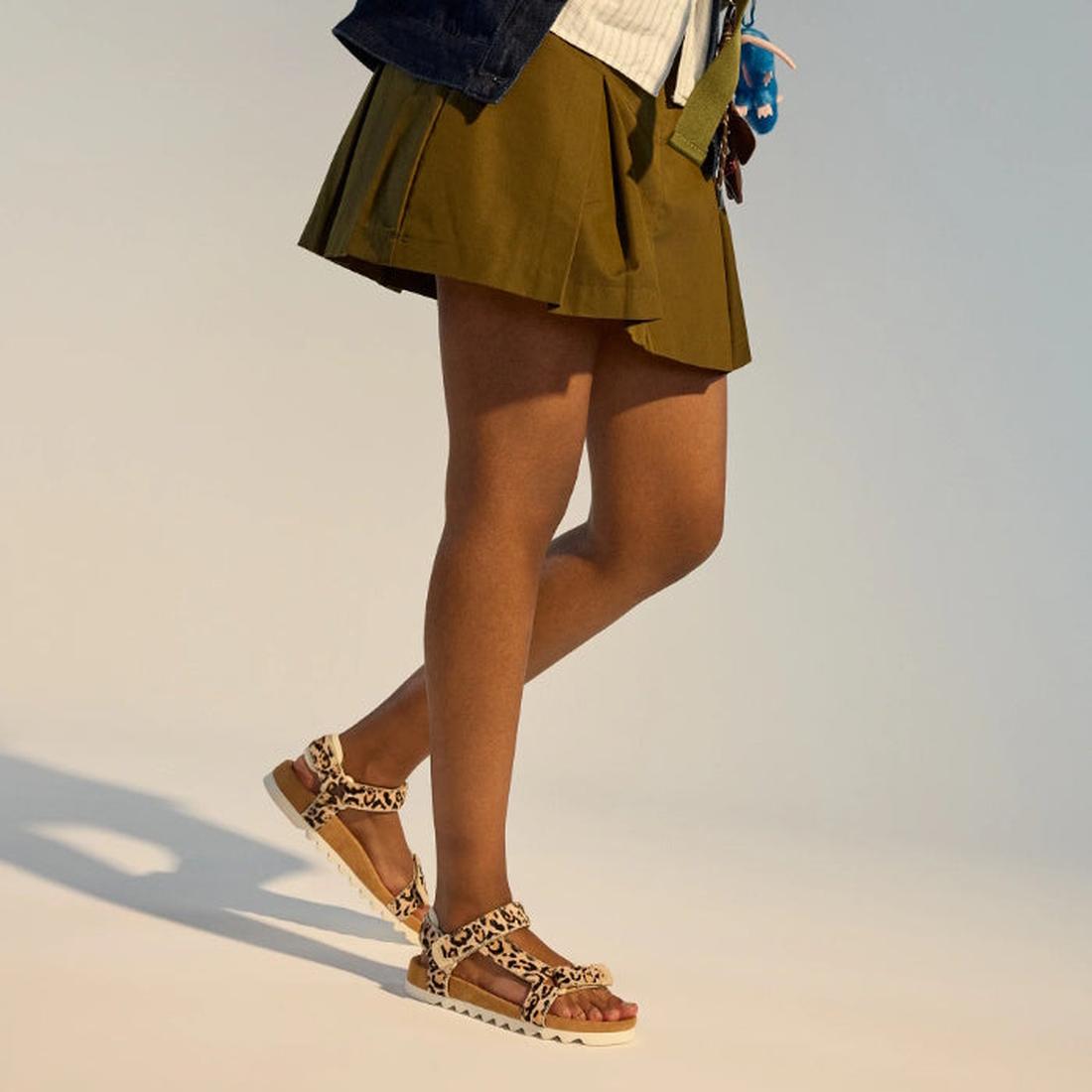 Wearing a short olive green skirt and Rollie Womens Sandal Tooth Wedge Neutral Leopard sandals with adjustable leather straps, a person stands against a neutral background, showing only the lower half of their body., 9382771798722, 9382771798739, 93827717