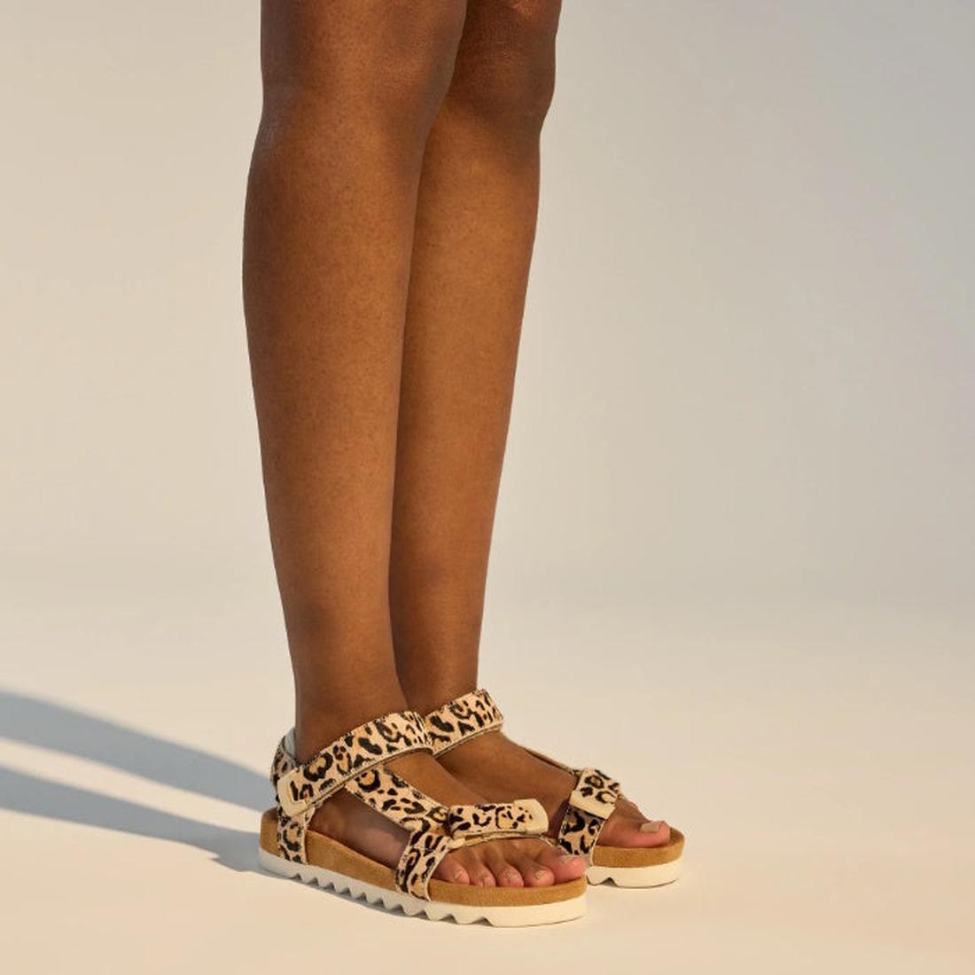 A person stands on a light surface, feet slightly apart, wearing the Rollie Womens Sandal Tooth Wedge in Neutral Leopard, featuring thick soles and adjustable leather straps. Only the lower legs and feet are visible., 9382771798722, 9382771798739, 9382771