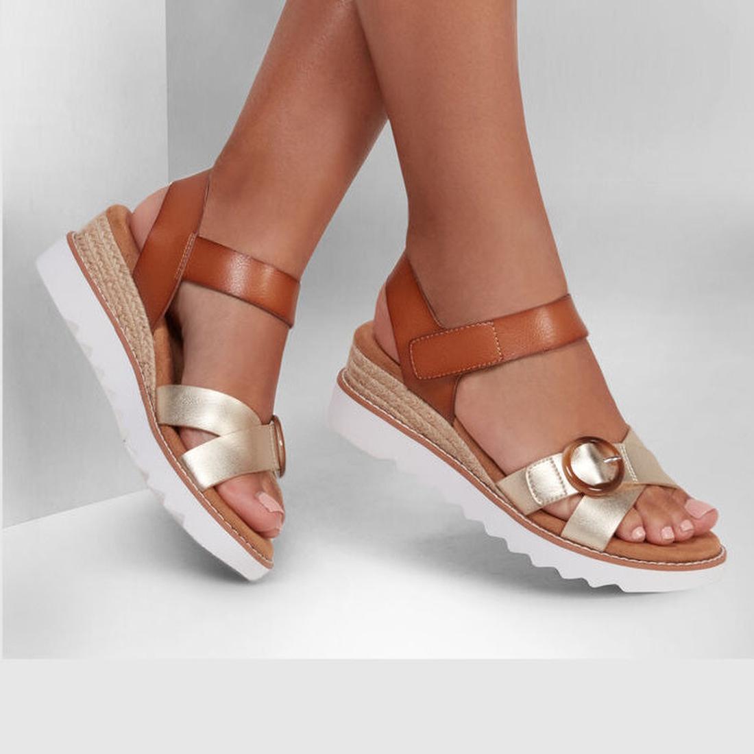 A person wears Skechers Bobs Womens Desert Kiss Hi Divine Garden Champagne Tan sandals with tan and metallic gold straps, a decorative buckle, and white platform soles, standing against a light background., 195969581407, 40918, 195969581414, 40920, 195969